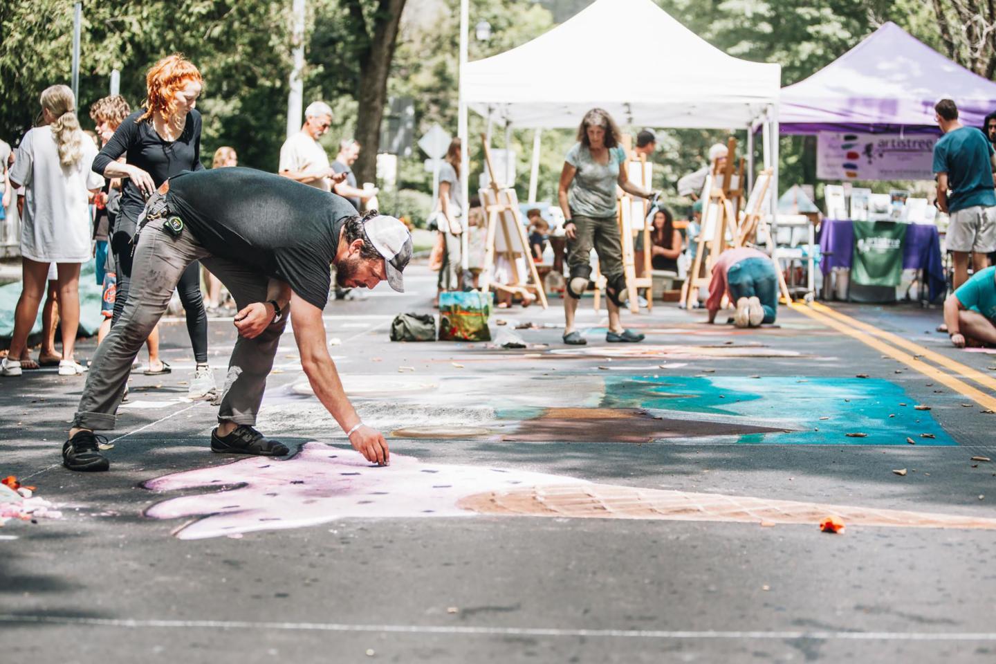 Man drawing with chalk while a crowd at the festival watches him. Tents are visable in the background as well as other people drawing and enjoying the festival.