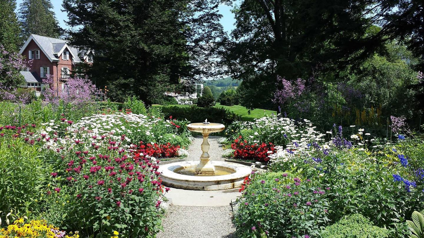 Garden with flowers in bloom. Fountain running in the middle of the flowers and the mansion is peeking through the trees in the back.