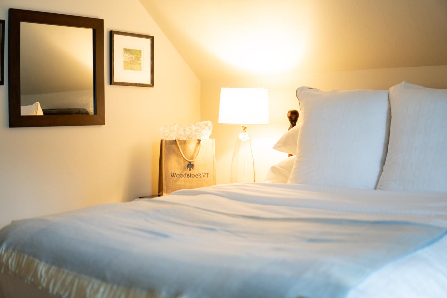 Queen bed with white pillows and sheets. Bedside lamp turned on with a Woodstock Inn gift bag next to the lamp.