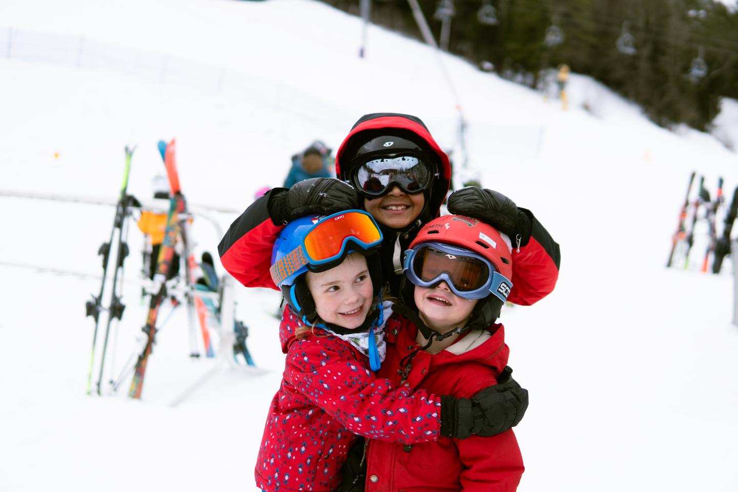 Three kids hugging with a ski area behind them