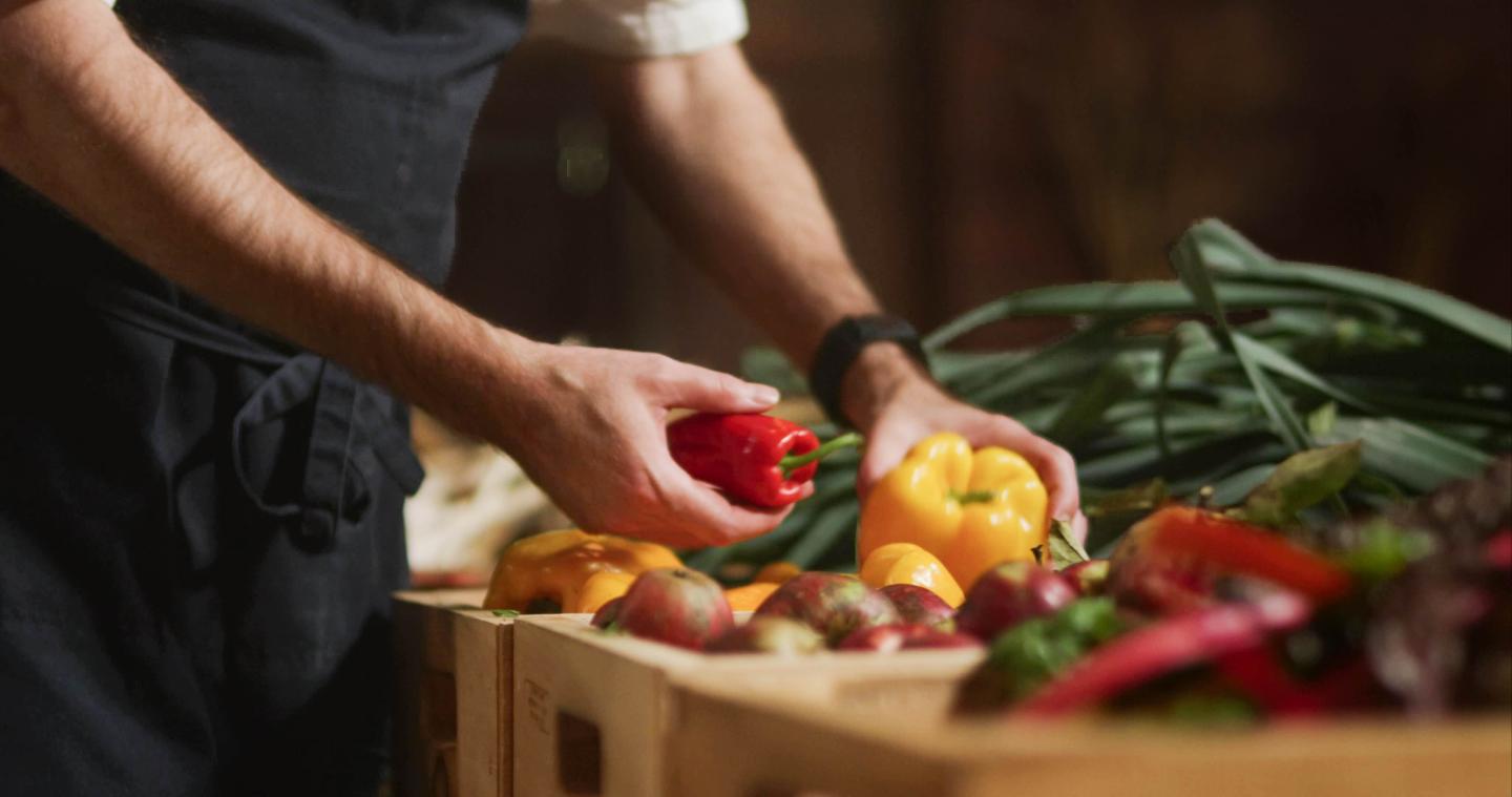 Chef picking up red and yellow bell peppers with baskets of produce in the background
