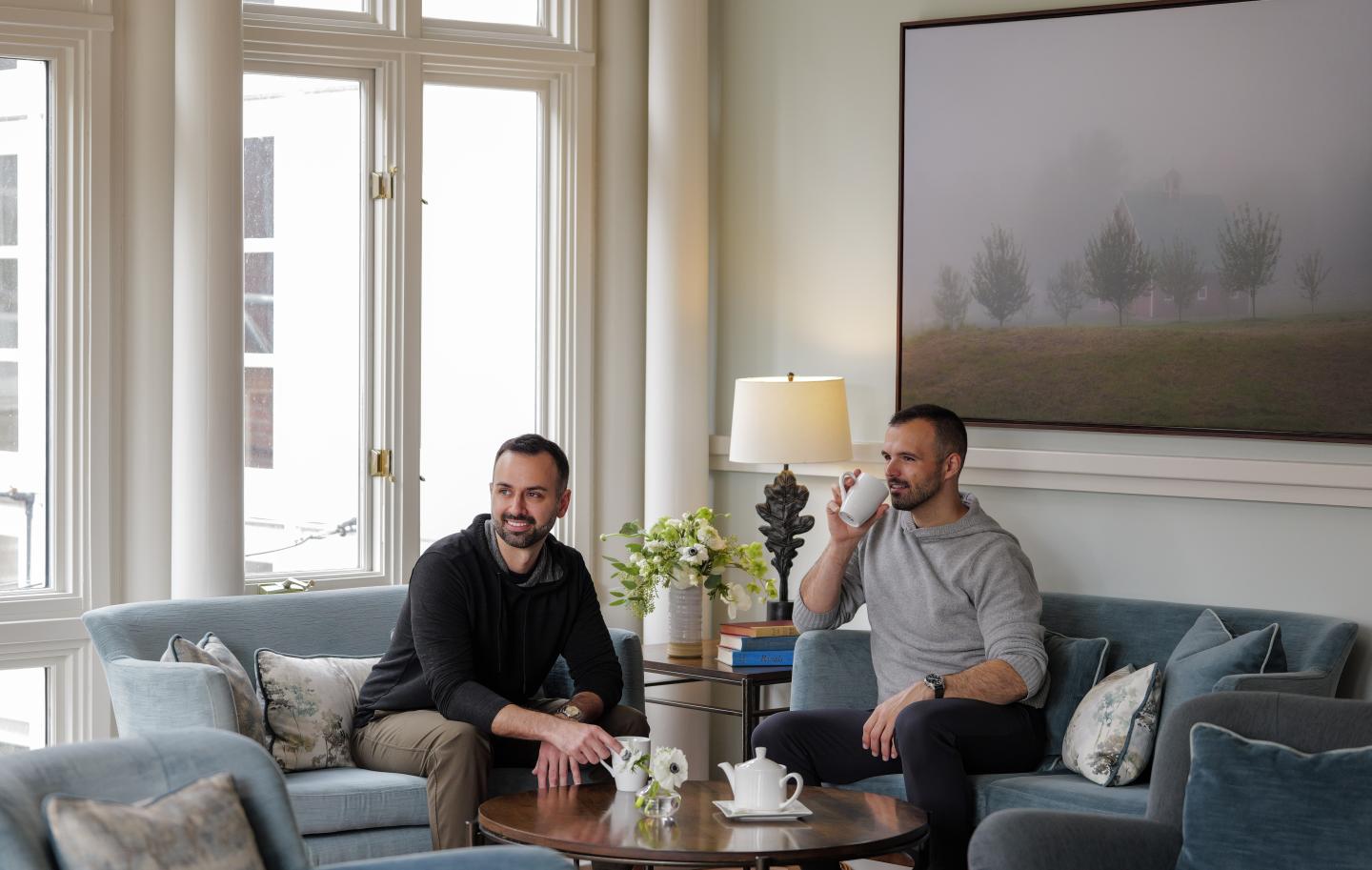 Two men sit on a blue sofa, smiling, in a bright living room.