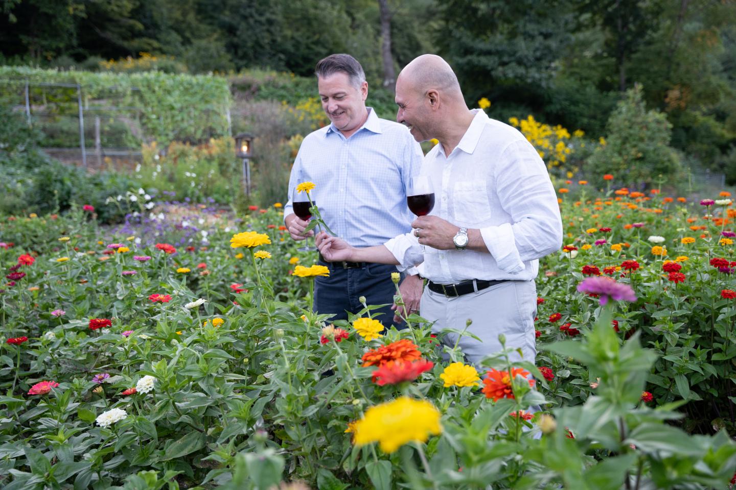 Two men with wine glasses in a colorful flower garden, smiling.