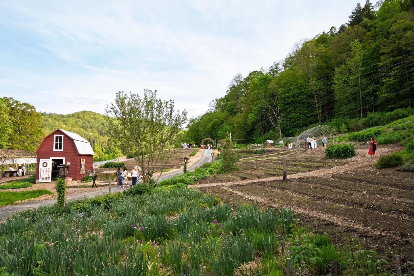 Red barn and people on a lush farm with garden plots and forested hills.