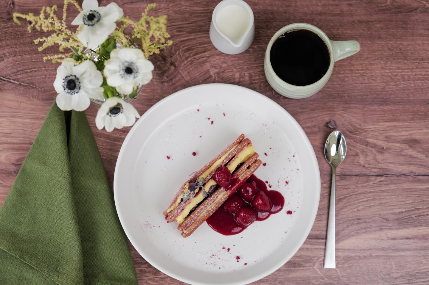 Slice of layered dessert with berries on a plate, coffee mug, flowers, and a green napkin.