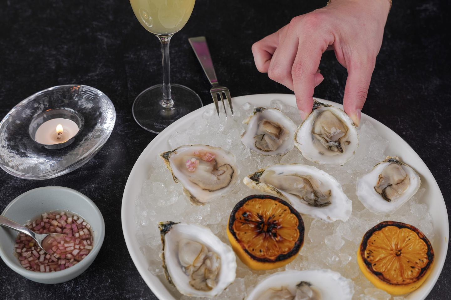 Oysters on ice with grilled lemons and a hand reaching for one.