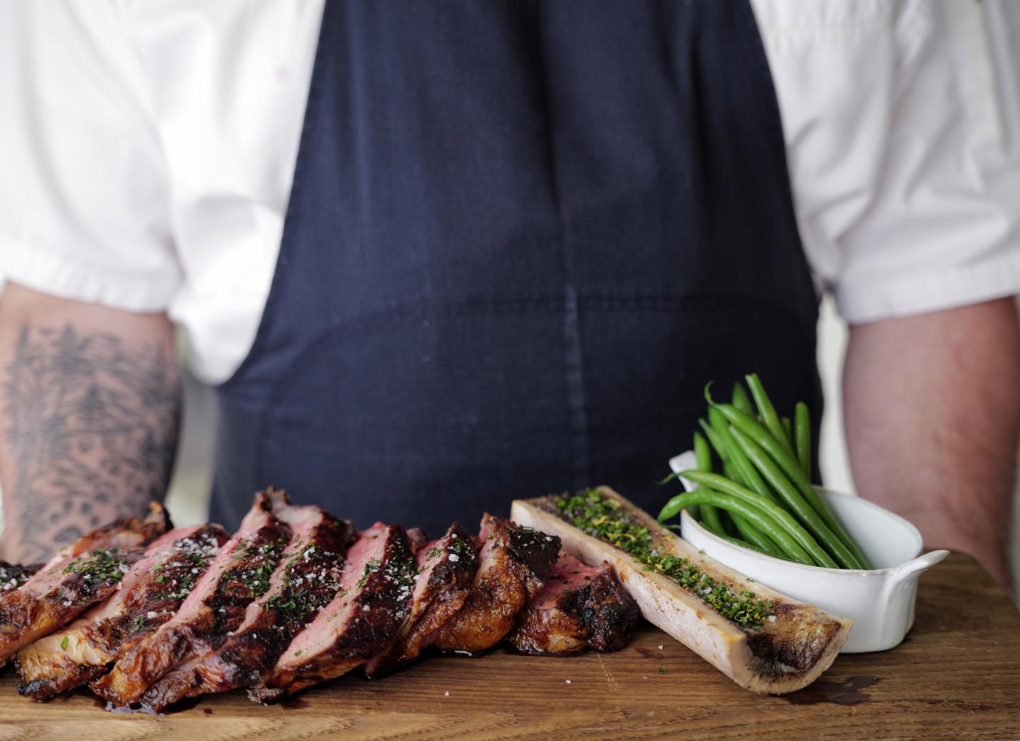 Chef in apron with sliced steak, green beans, and garnish on wooden board.