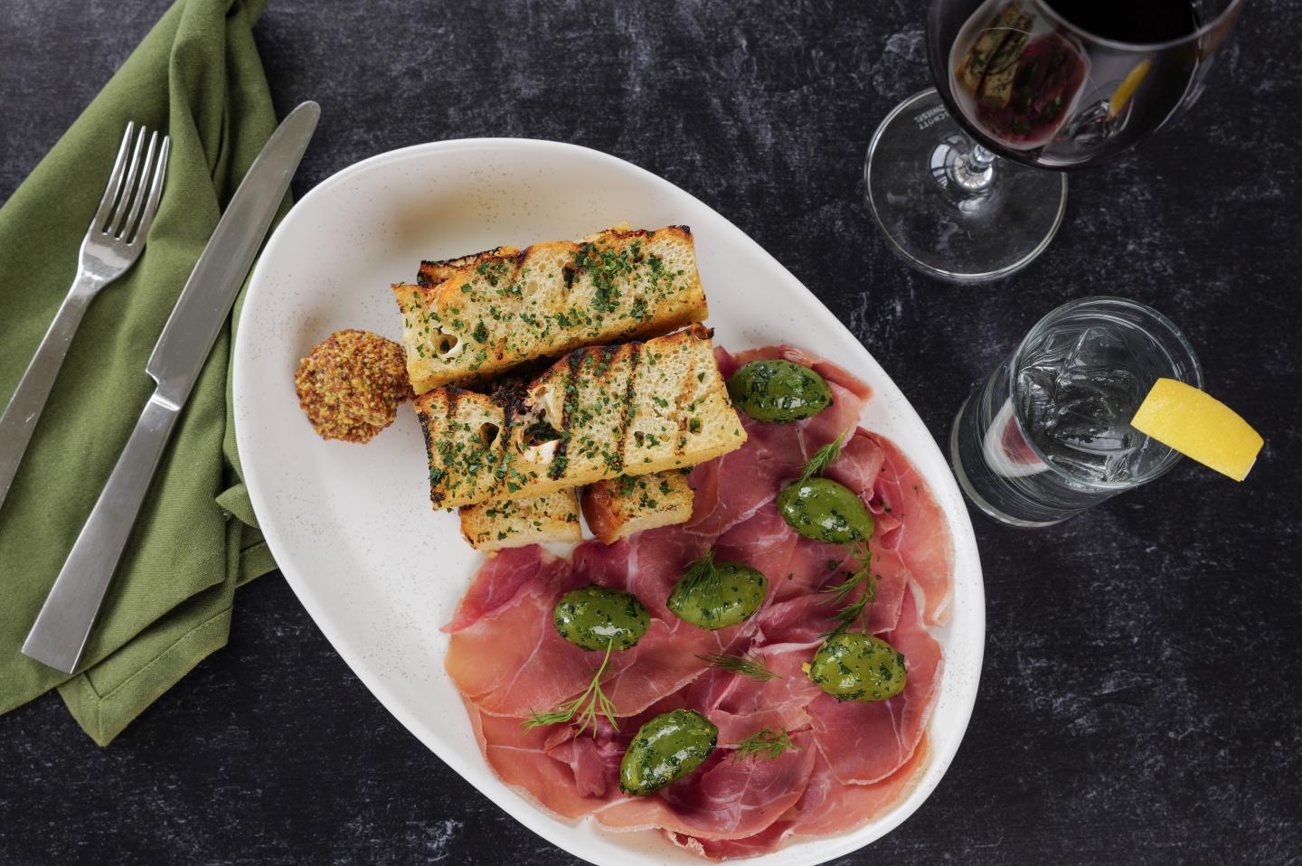 Cured ham and olives with herb bread on a white plate, beside wine and water glasses.