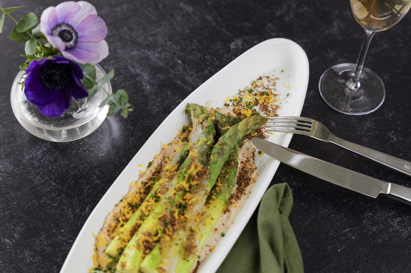 Asparagus with grated topping on a white plate, next to a flower vase and wine glass.