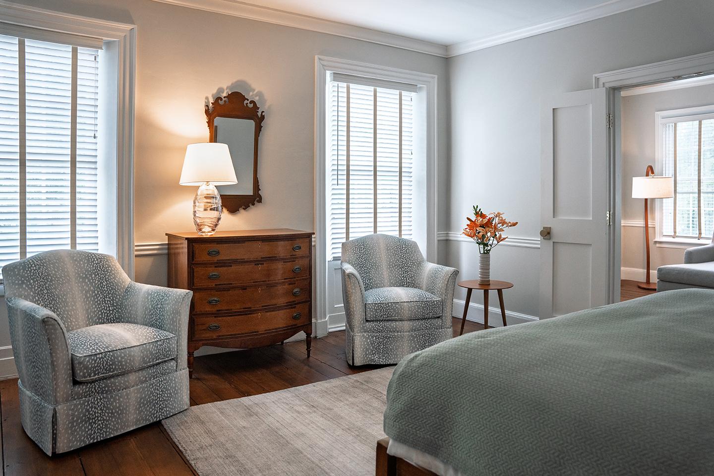 Cozy bedroom with two armchairs, a dresser, and a bed near large windows.