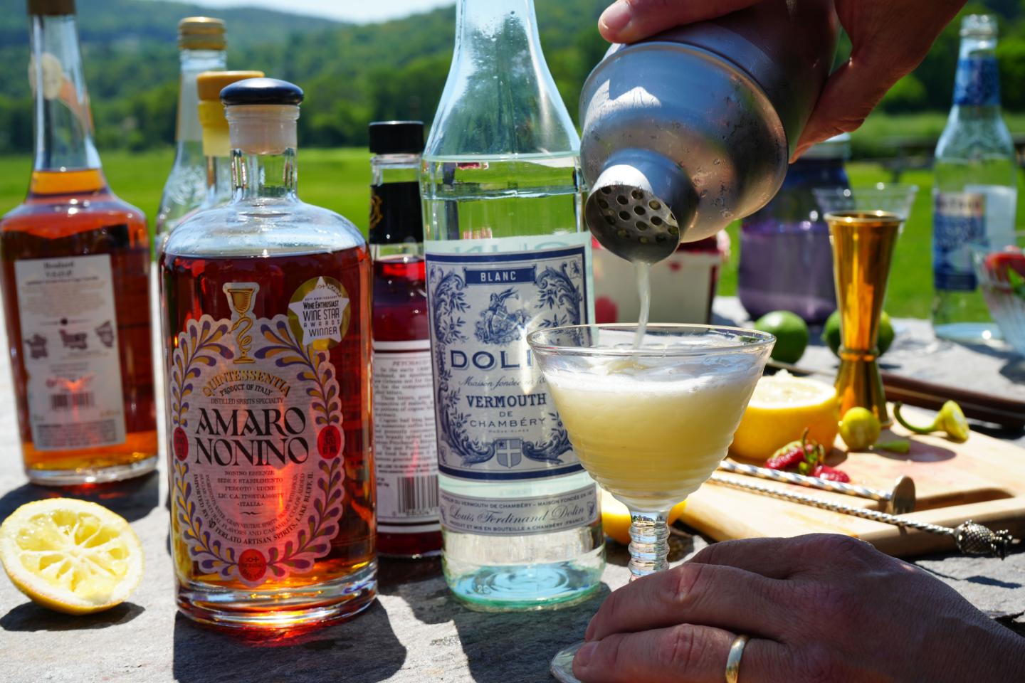 Mixologist pouring cocktail in sunny outdoor setting. Bottles and sliced lemon on table.