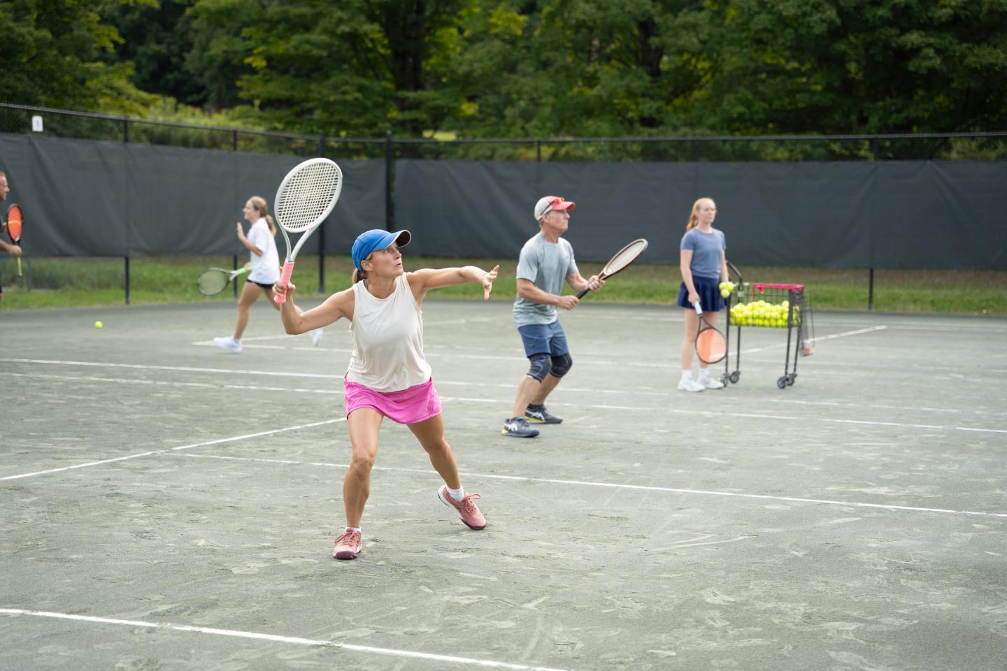 Tennis players practicing on a clay court, one woman mid-serve.