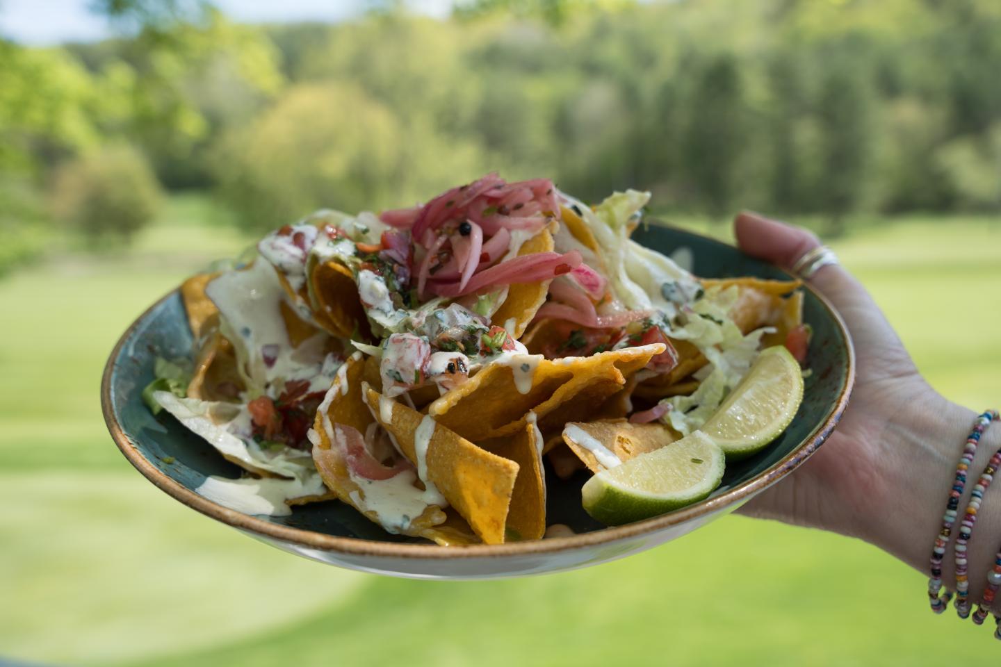 Plate of nachos with toppings, held outdoors with a grassy background.