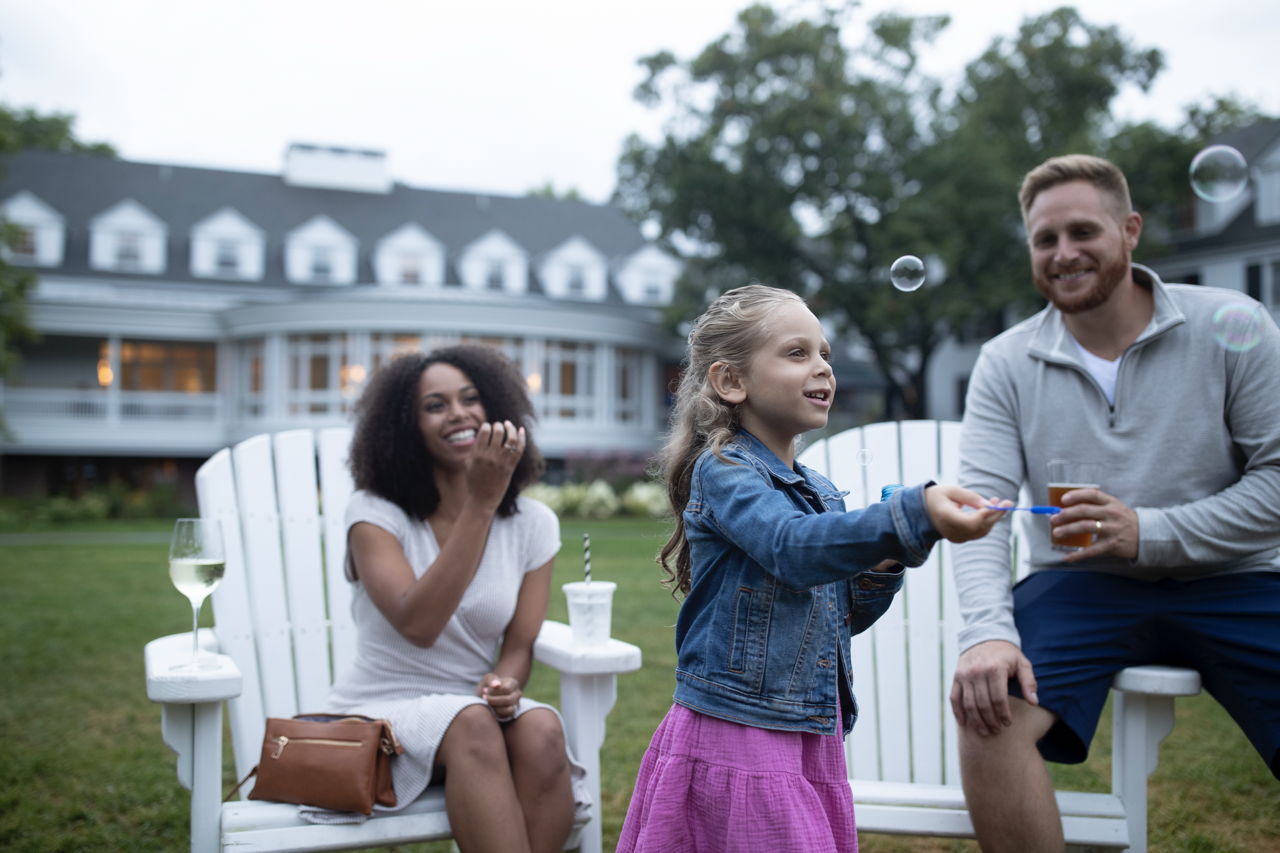 Child playing with bubbles, seated adults smiling outdoors near a large house.