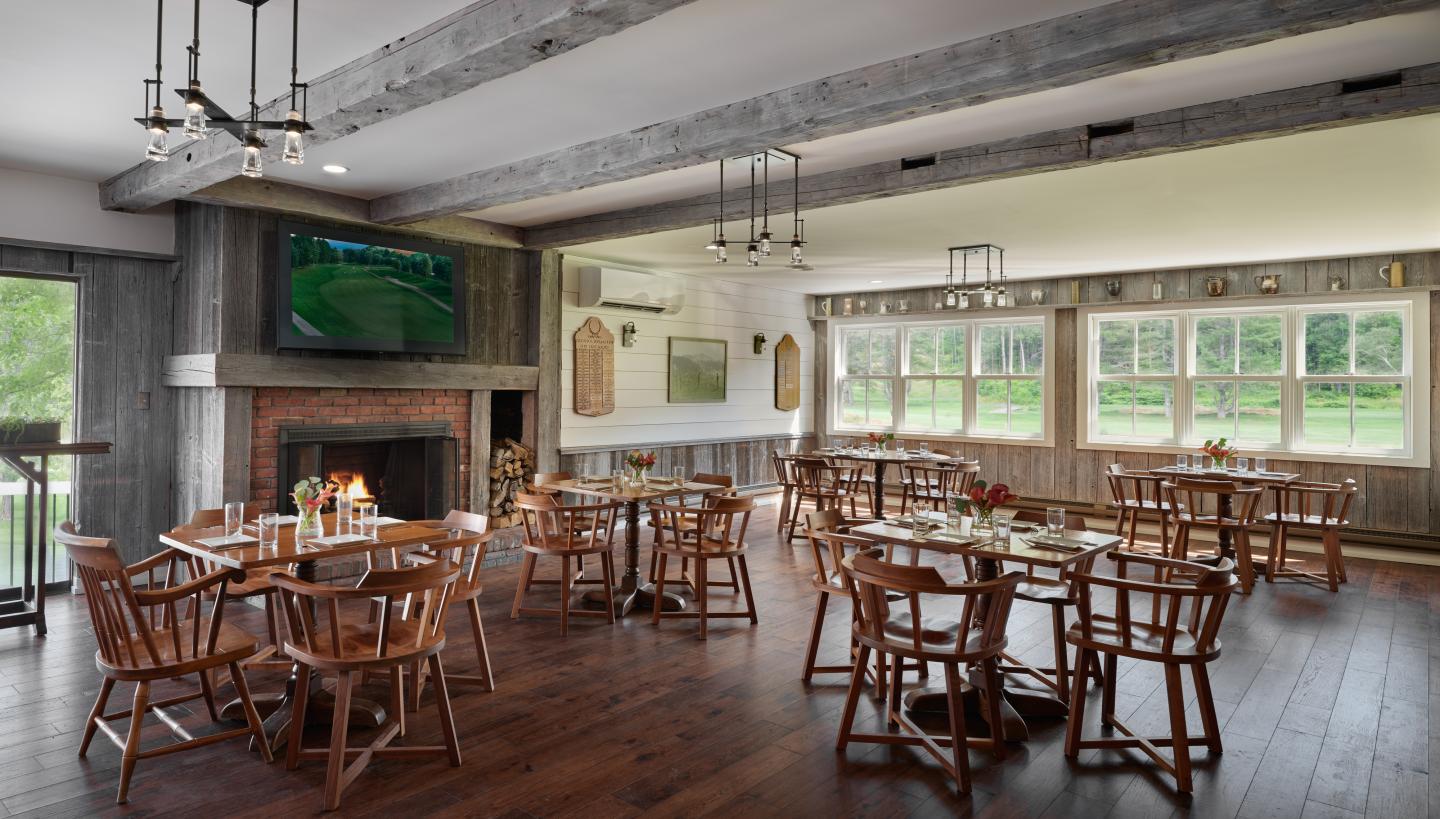 Spacious dining room with wooden tables and chairs, large windows, and a TV above the fireplace.