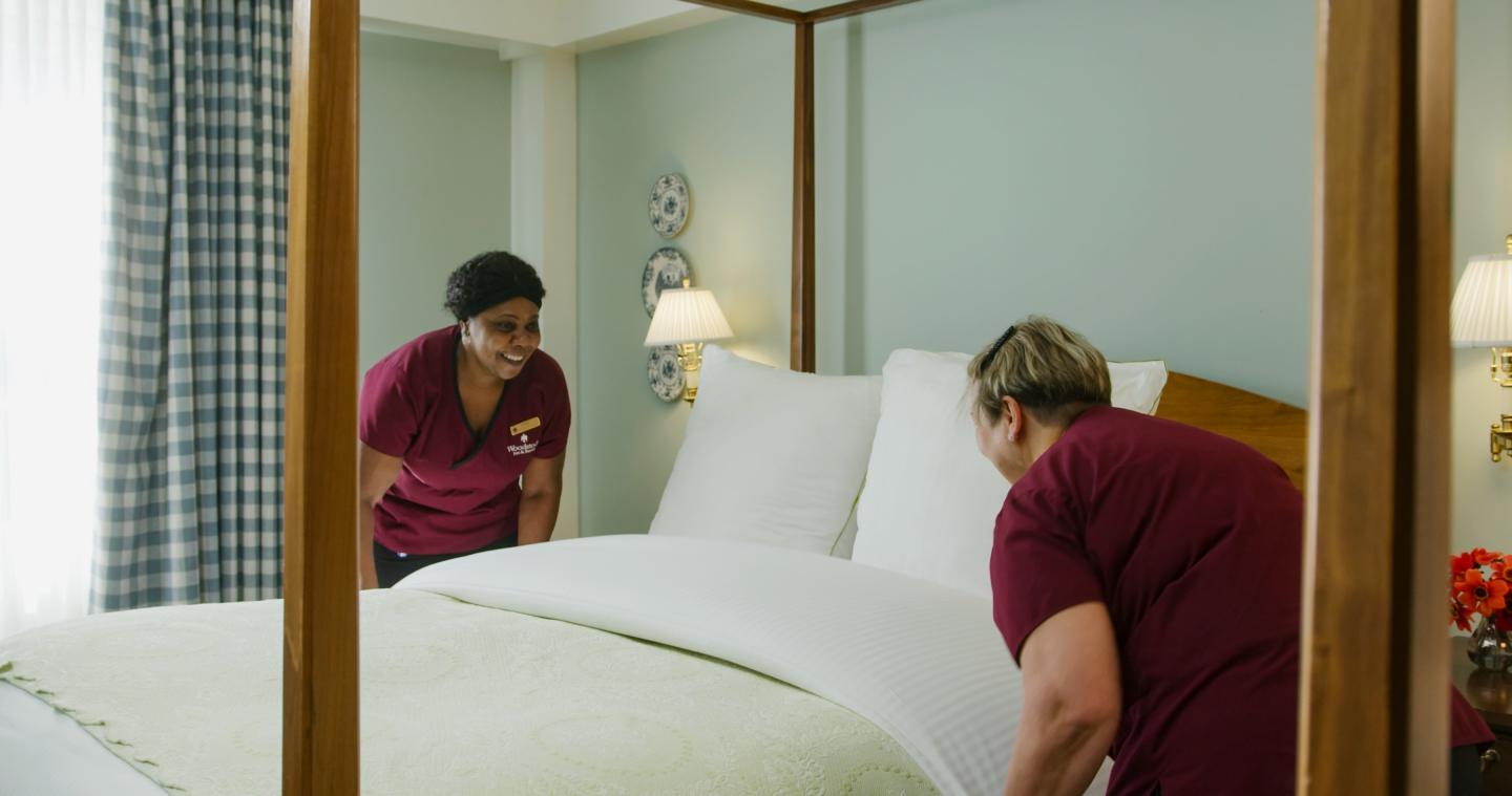 Two workers in maroon shirts making a bed in a hotel room.