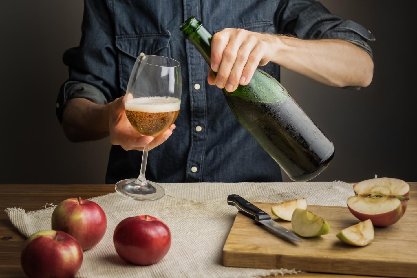 Pouring cider into a glass on a wooden table with apples and sliced apples on a cutting board.