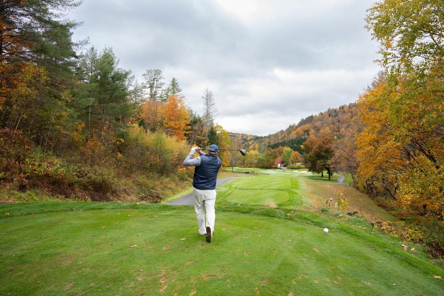 Golfer swings on a lush course surrounded by autumn trees under a cloudy sky.