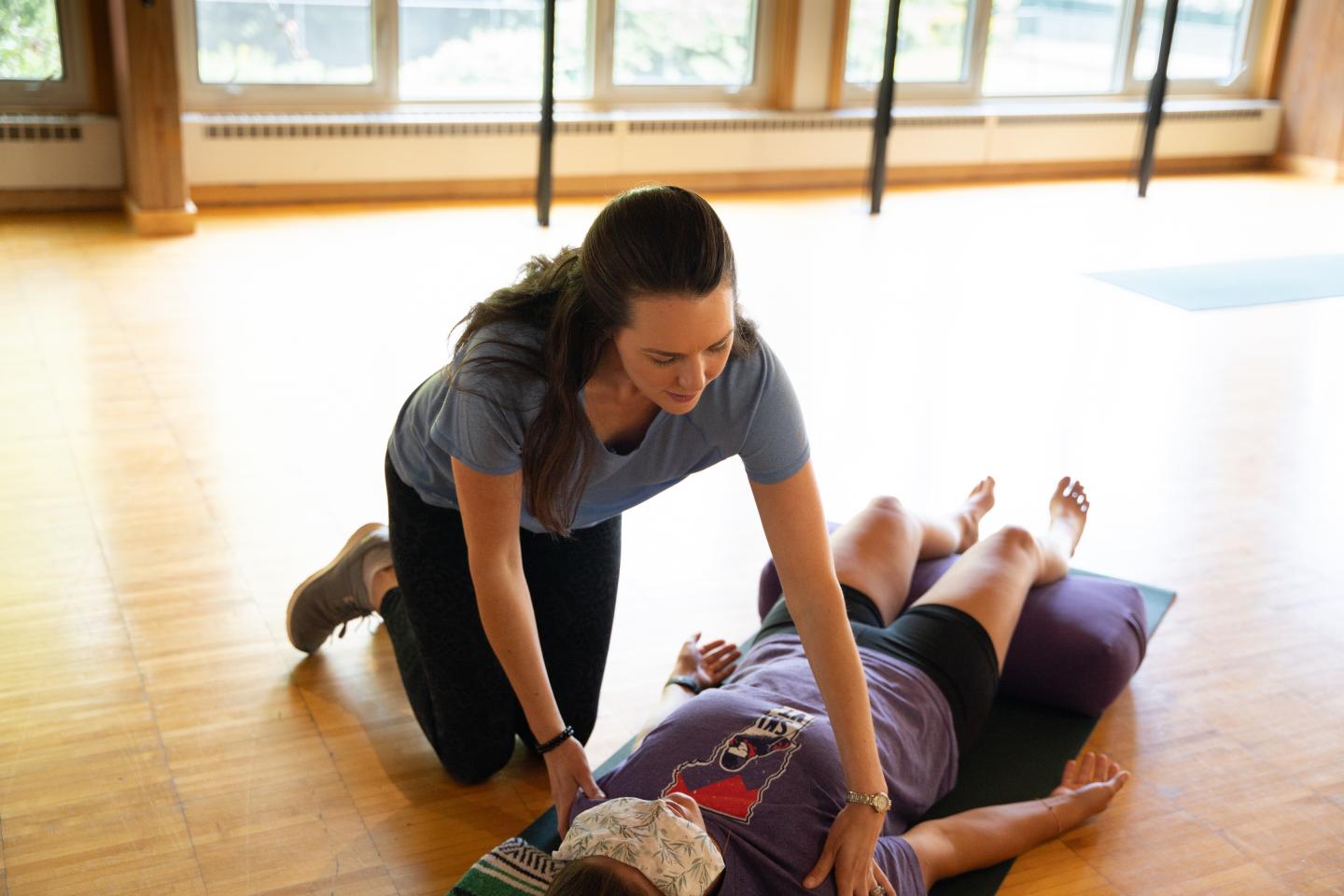 Yoga instructor assists student lying on mat in bright studio.