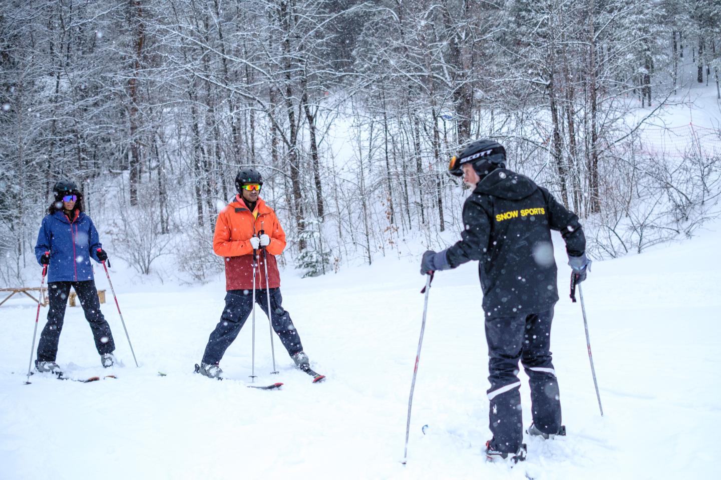 Three people in ski gear standing in a snowy forest, talking and holding ski poles.