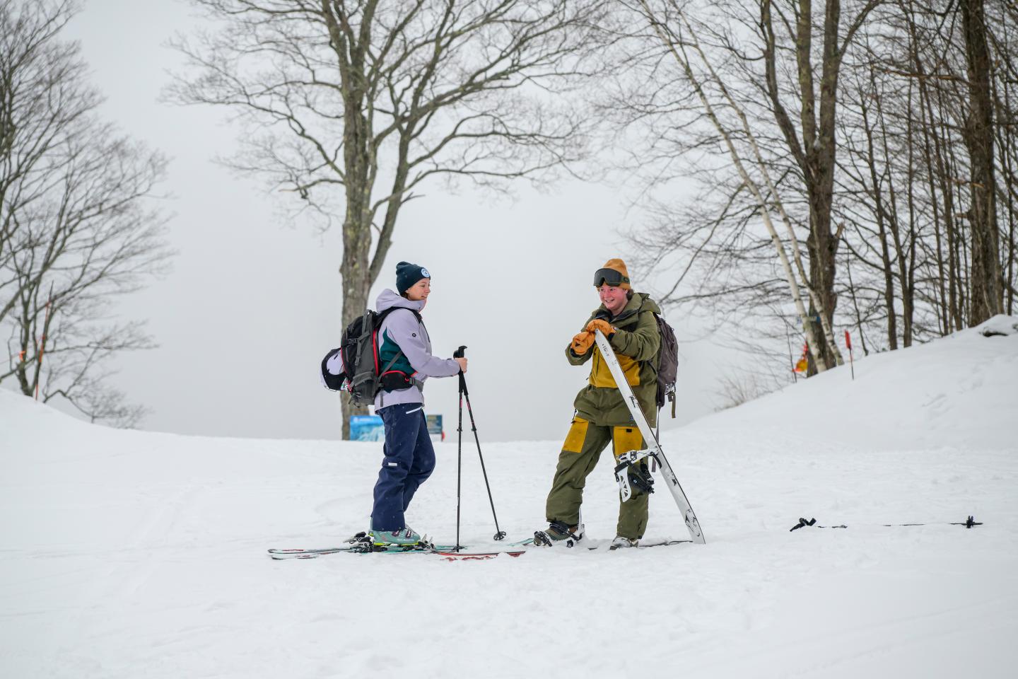 Two people in ski gear talking on a snowy slope with trees.