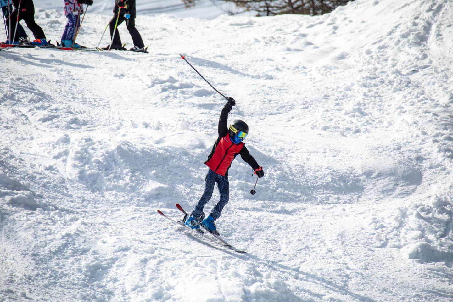 Skier in red jacket downhill on snowy slope, lifting one ski pole.