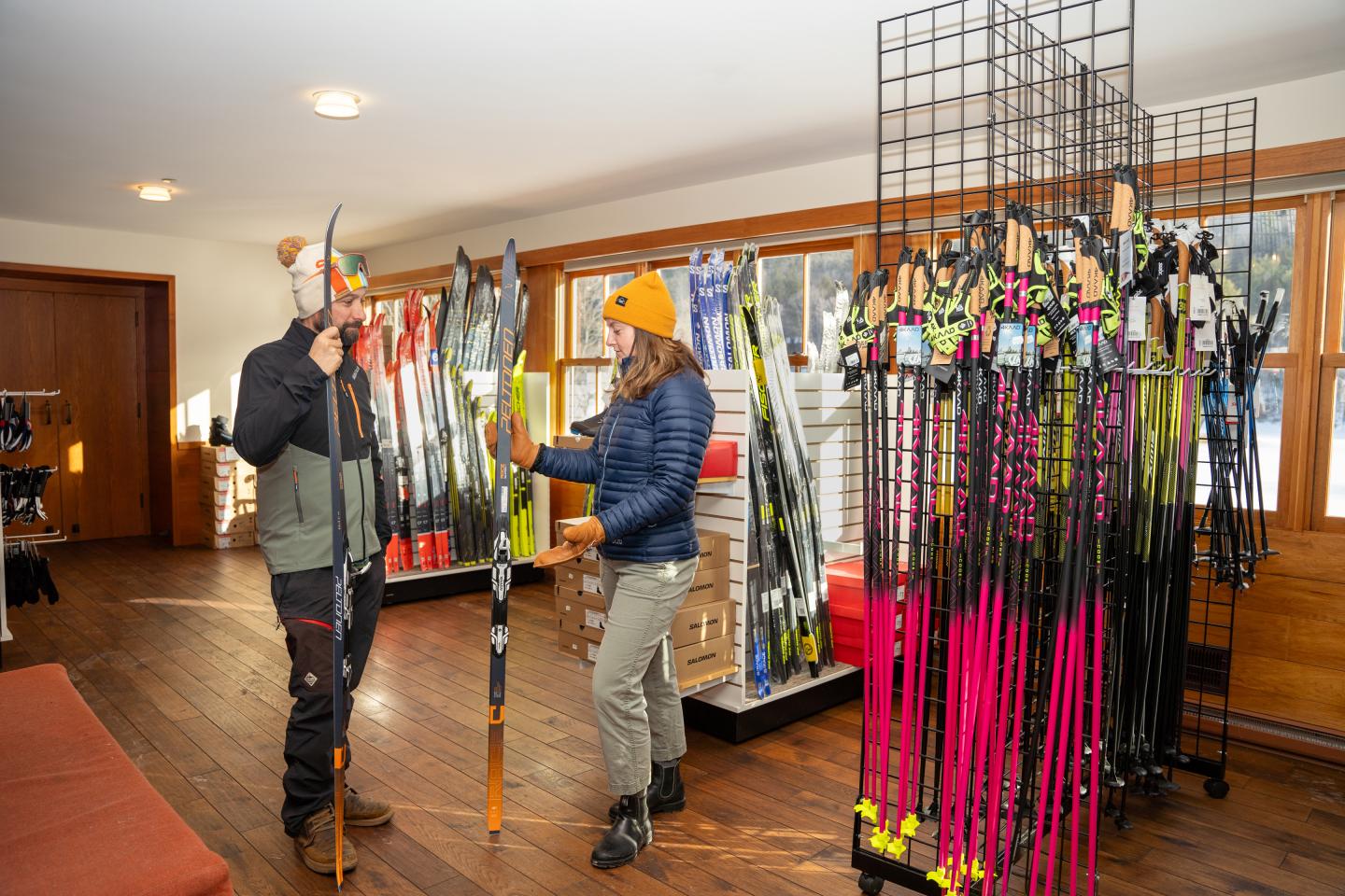 Two people choosing skis inside a shop with wooden floors and large windows.