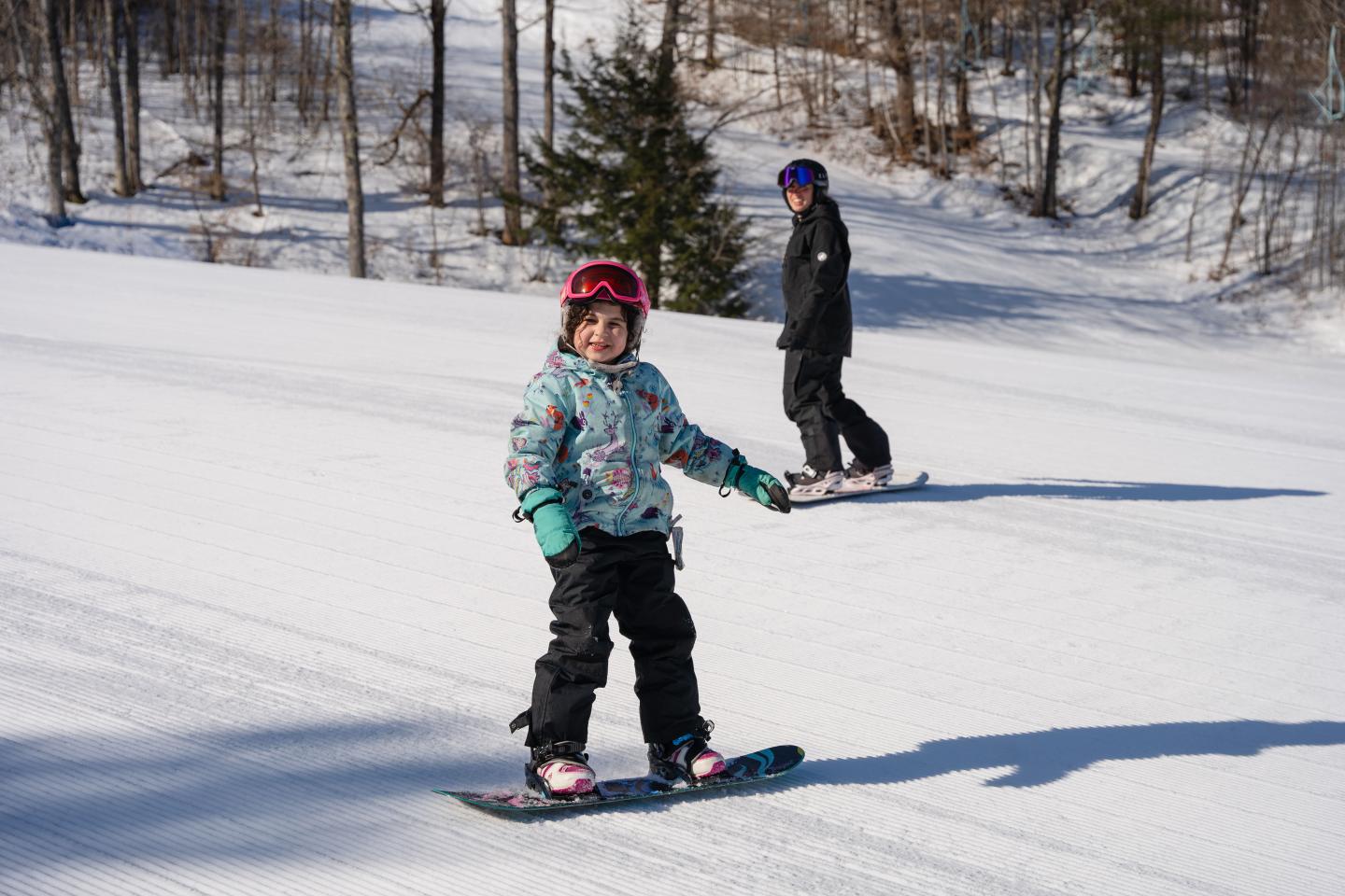 Child snowboarding on a snowy slope with an adult in the background.