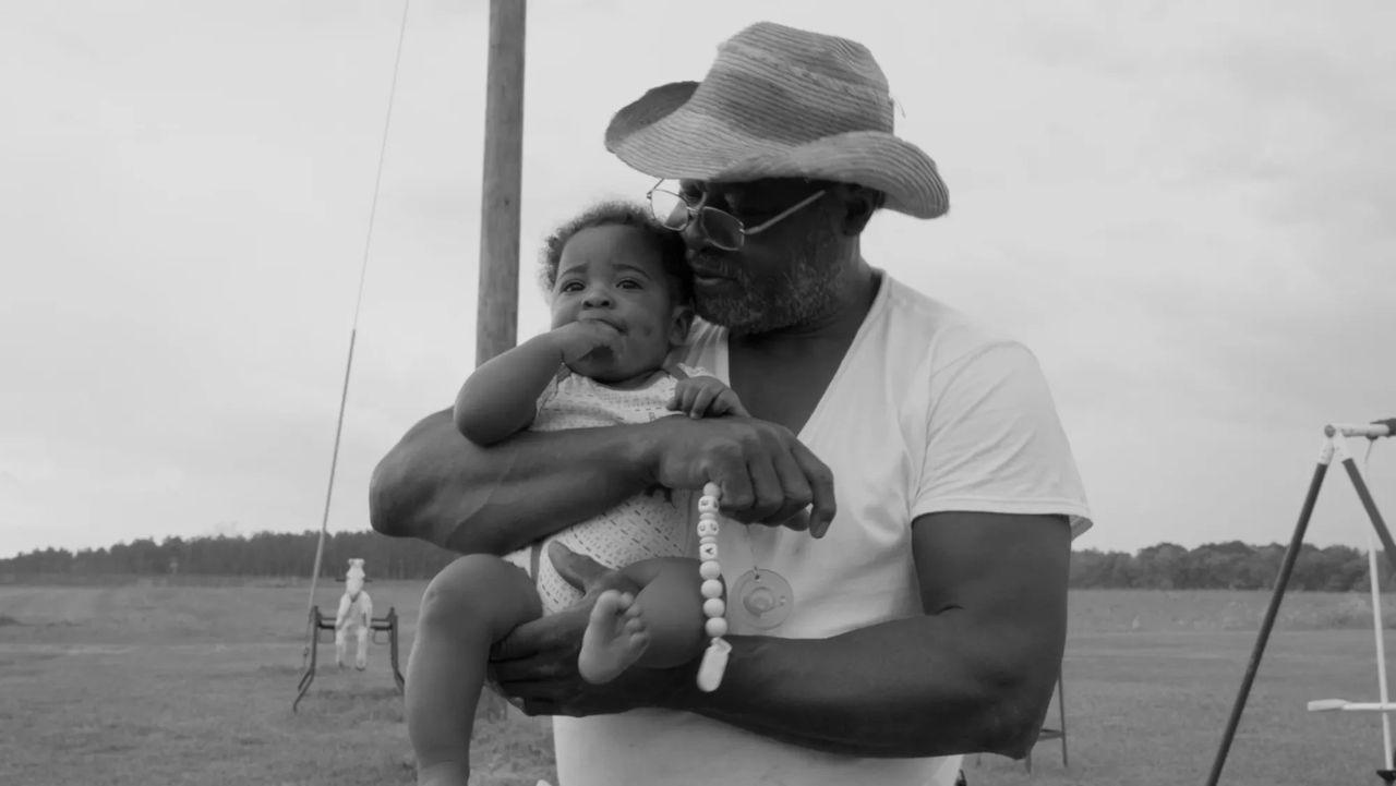 Elderly man holding a baby, both relaxed outdoors in grayscale.