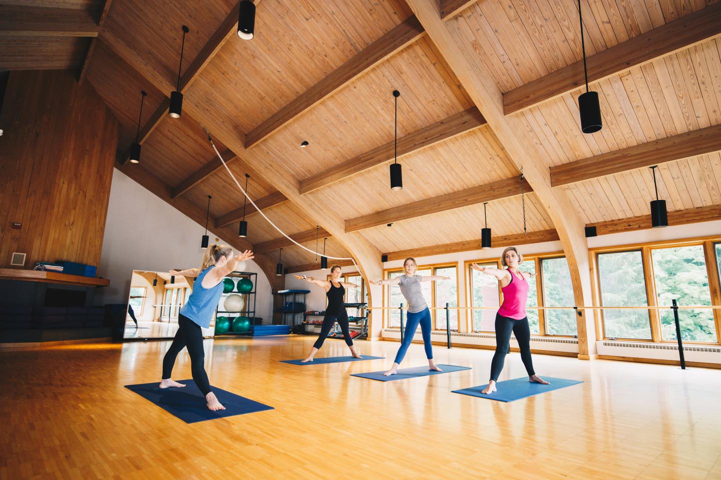 Yoga class in a spacious wooden hall with people on mats stretching.
