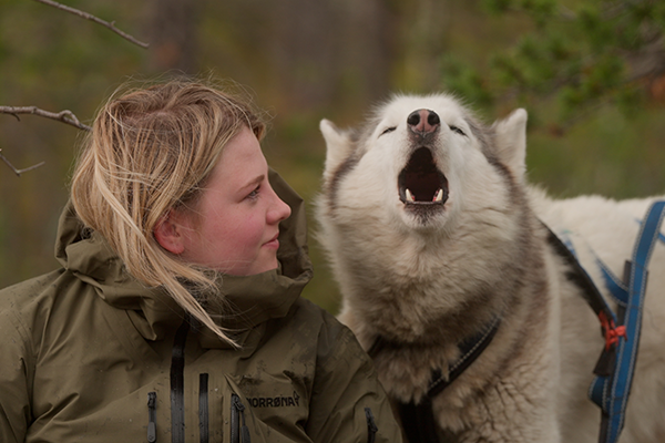 Woman in a green jacket with a howling husky outdoors.