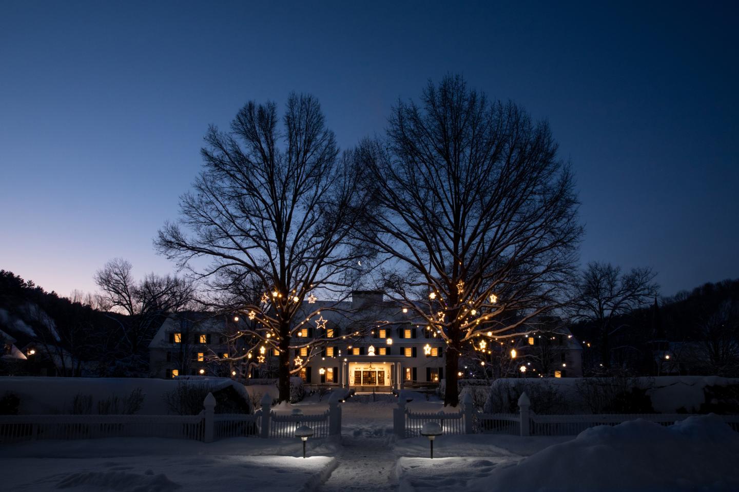 Snow-covered mansion at dusk, trees lit with warm lights.