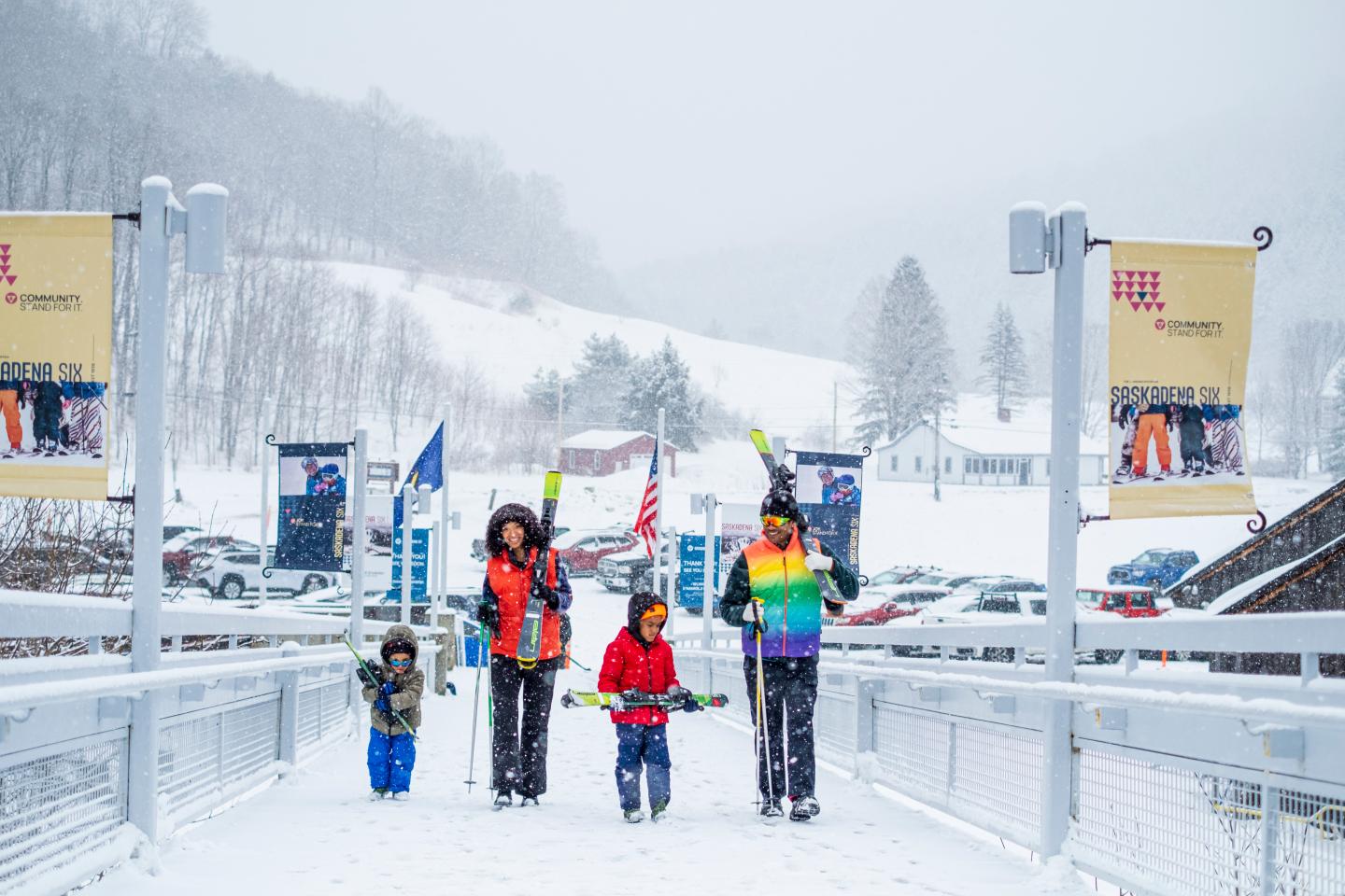 Family in winter clothing walking on snowy bridge at a ski resort.