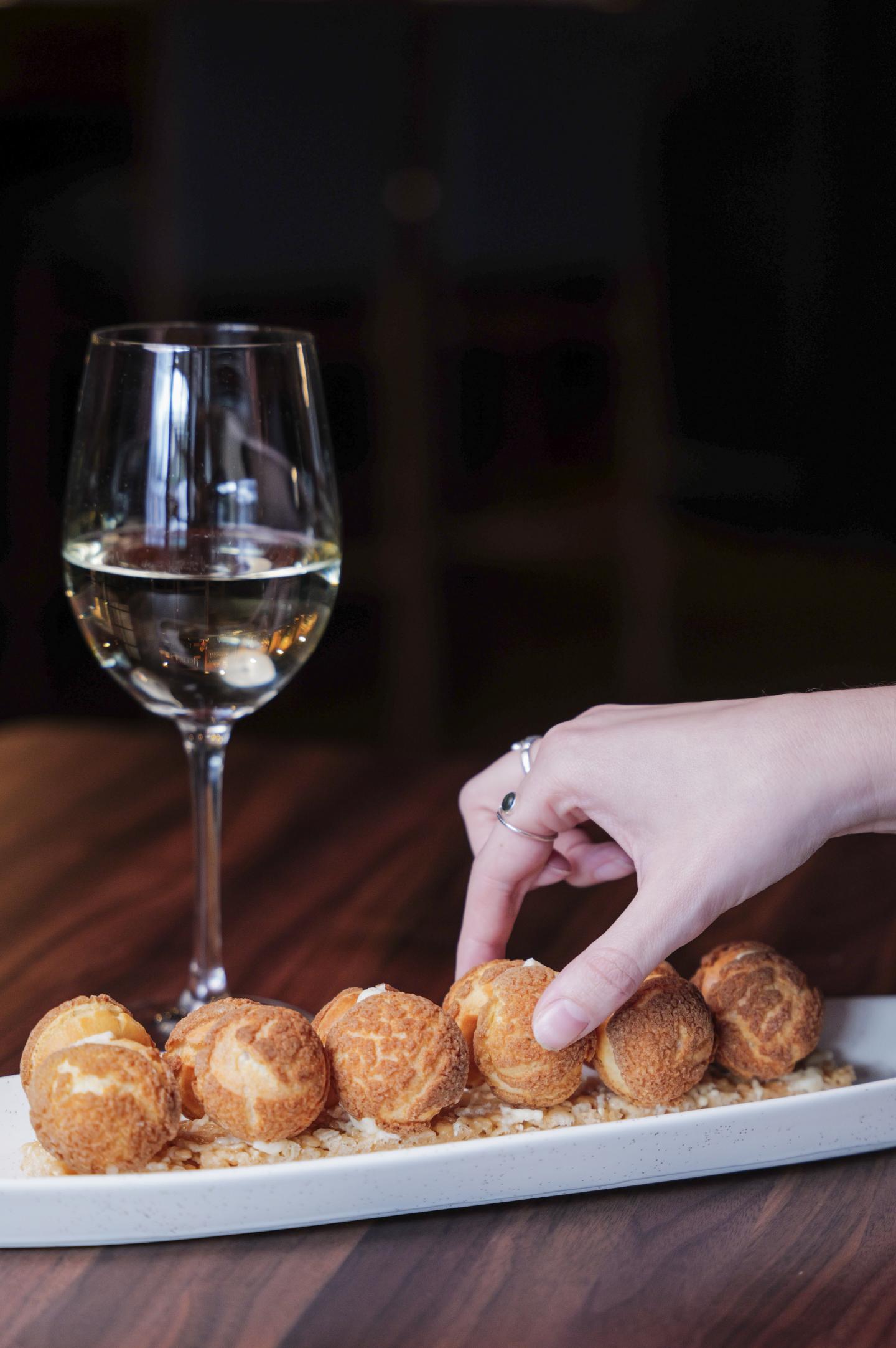 Hand reaching for a pastry near a glass of white wine on a wooden table.