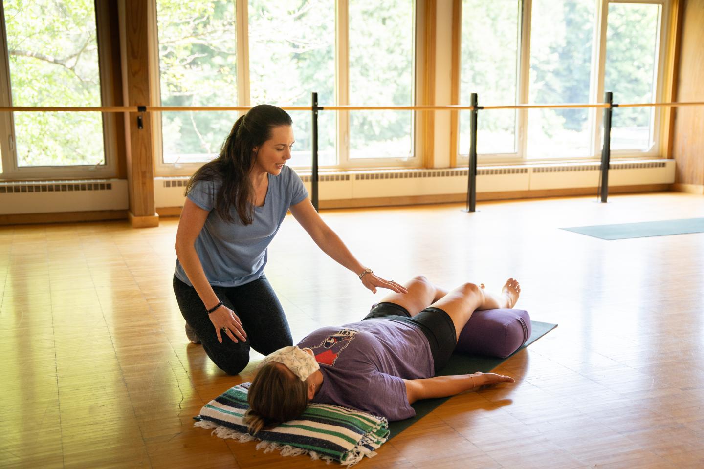 Yoga instructor guiding a person in a relaxation pose on a mat in a sunlit studio.