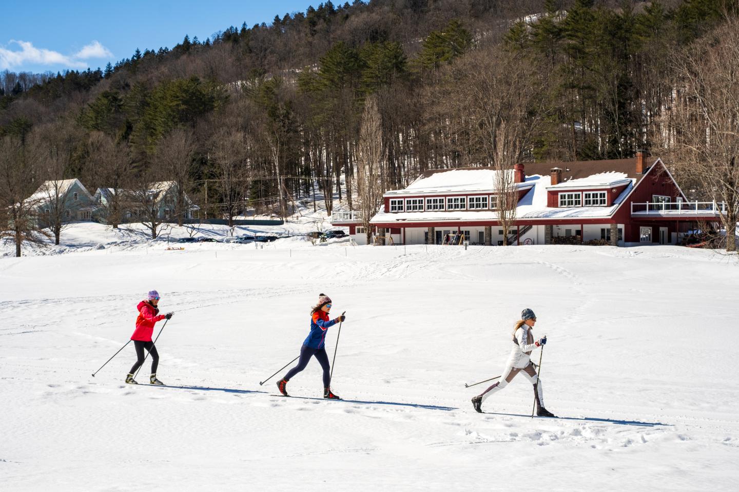 Three people skiing on snow near a lodge, trees in the background.