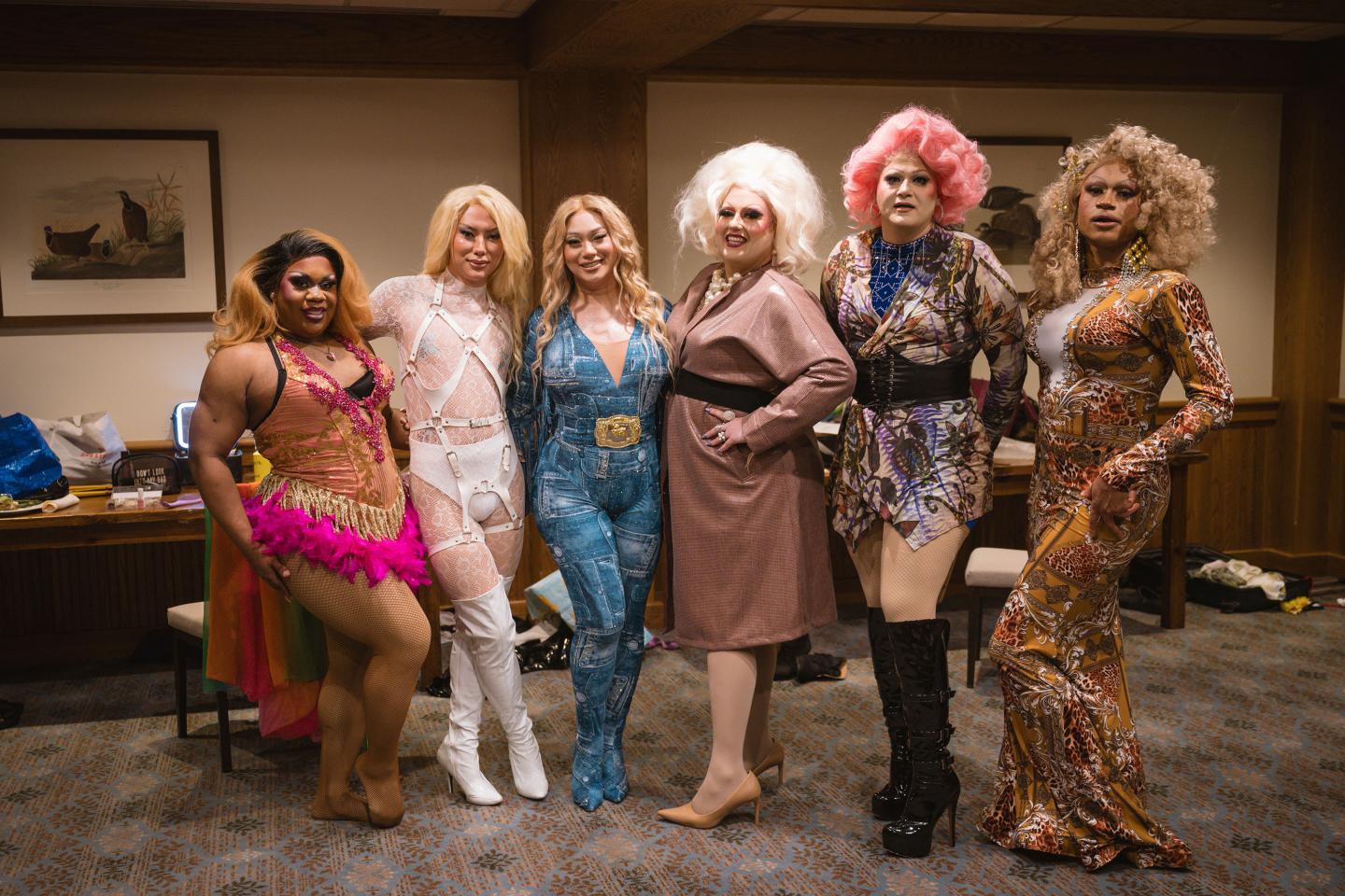 Drag performers in colorful outfits pose together in a wood-paneled room.