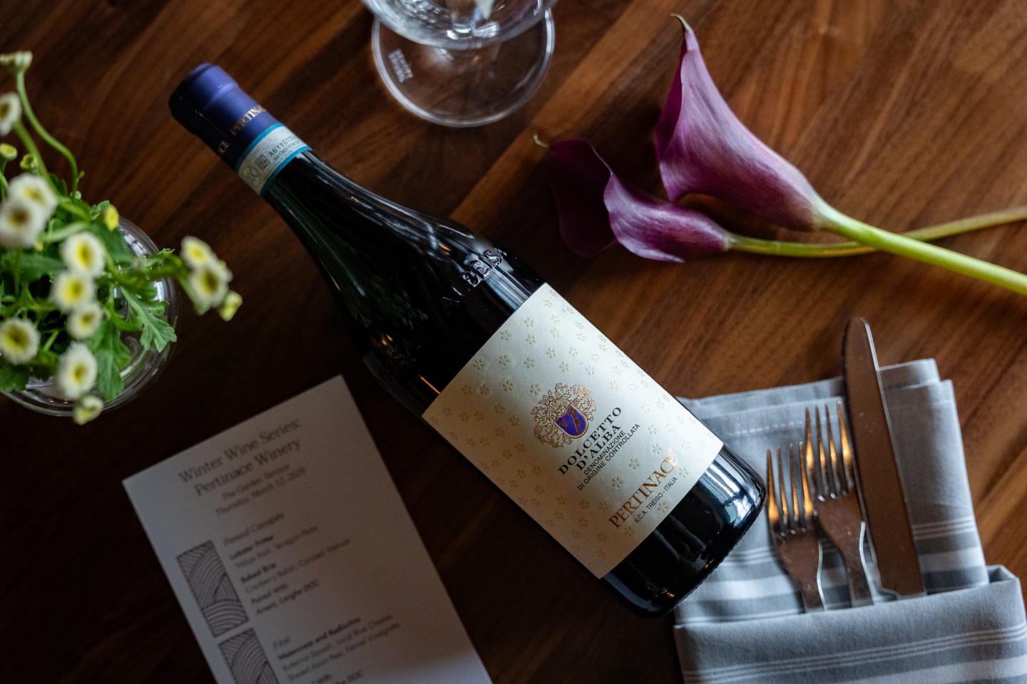 Wine bottle beside flowers, cutlery, menu, and glass on a wooden table.