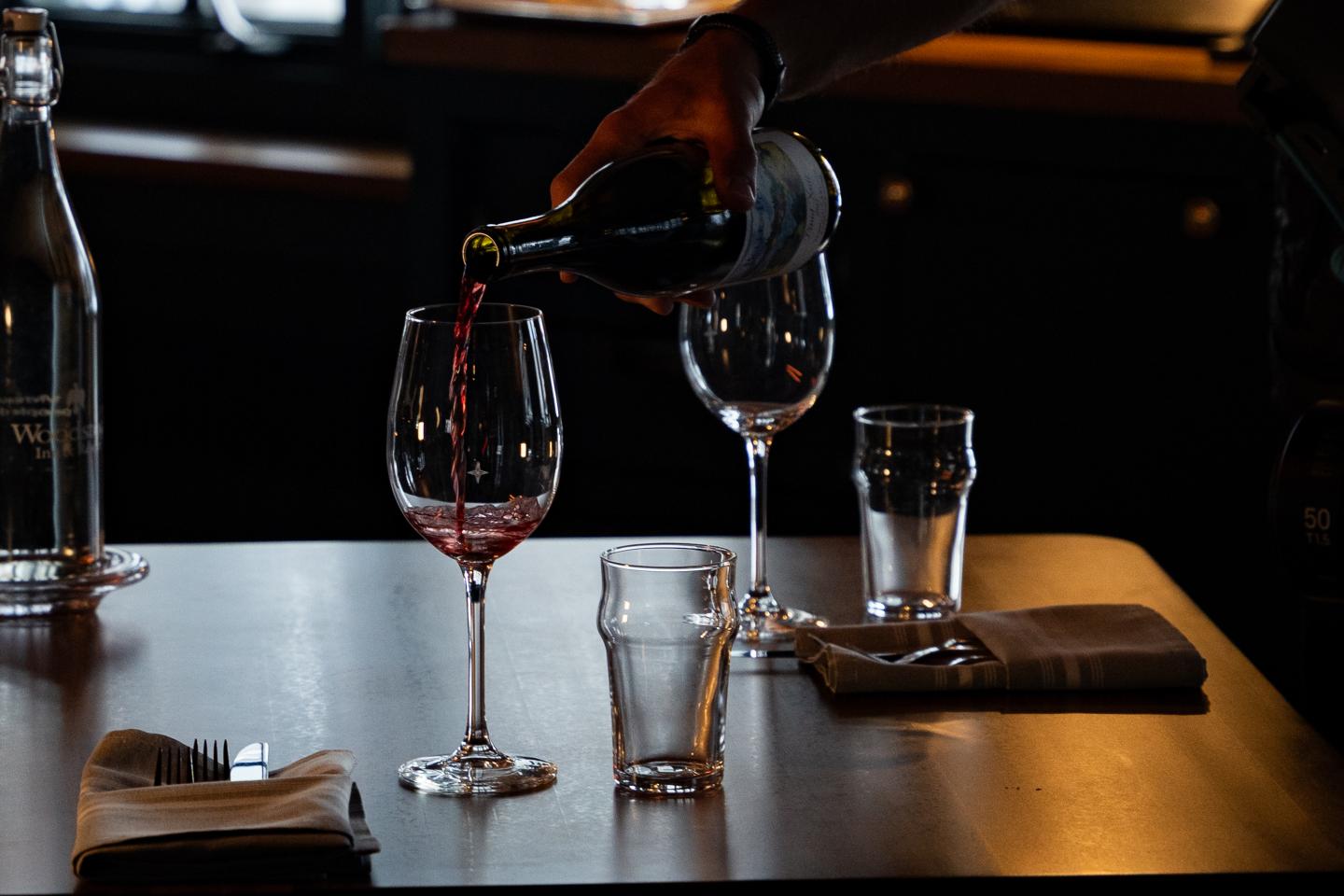 Pouring red wine into a glass on a dimly lit table with glasses and napkins.