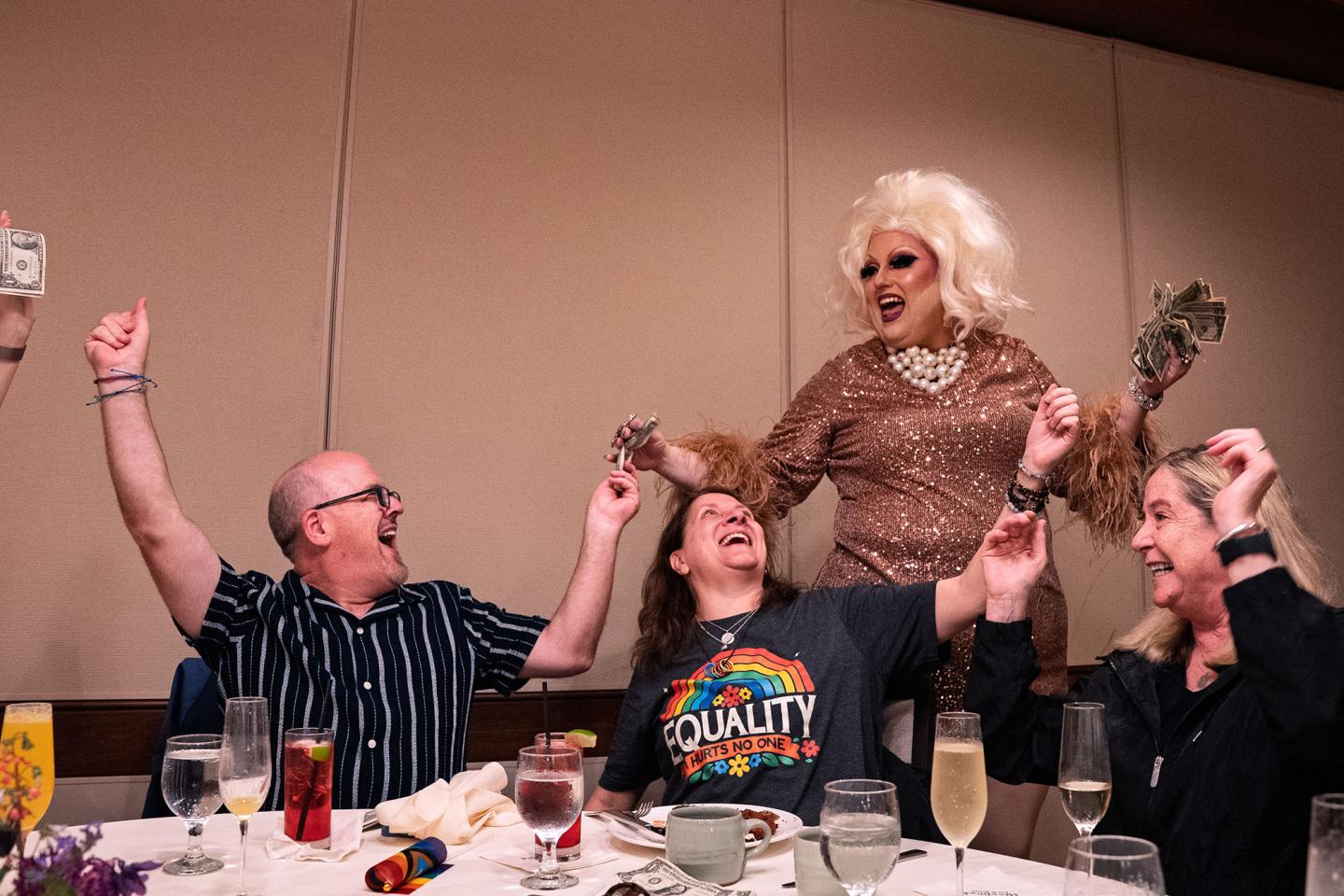 Drag performer with guests at a dinner table, all cheering joyfully.