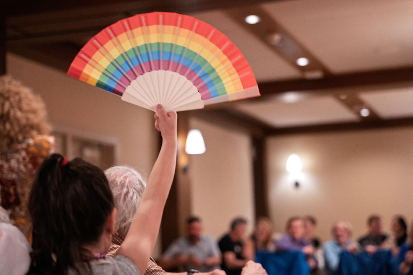 A person holds up a rainbow-colored fan in a room with seated people.
