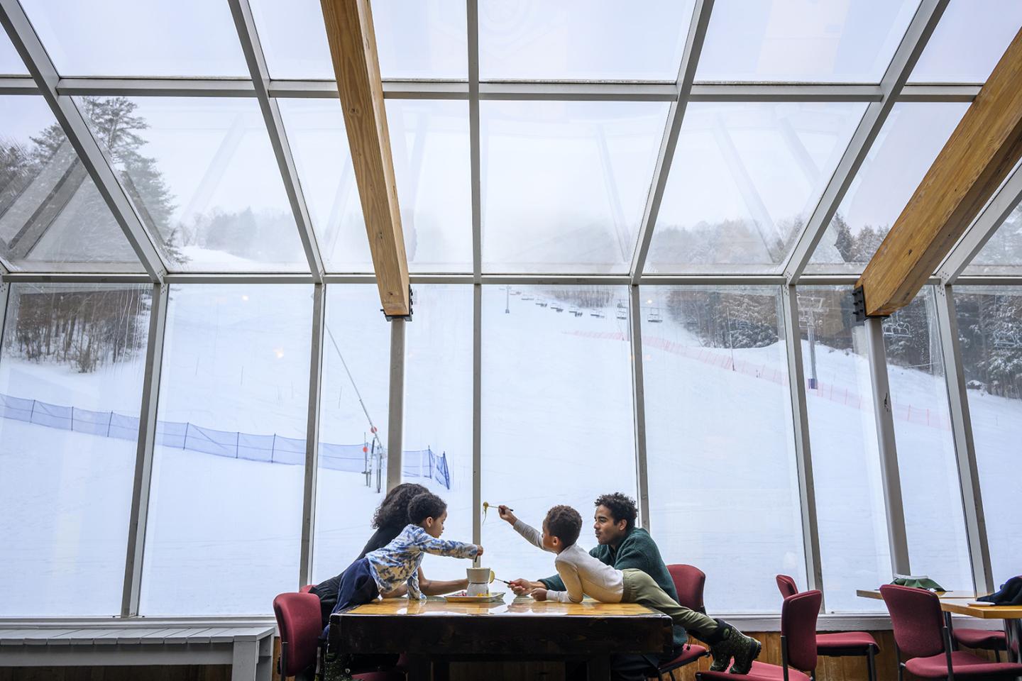 Family dining by large windows, snowy Saskadena 6 ski slope outside.