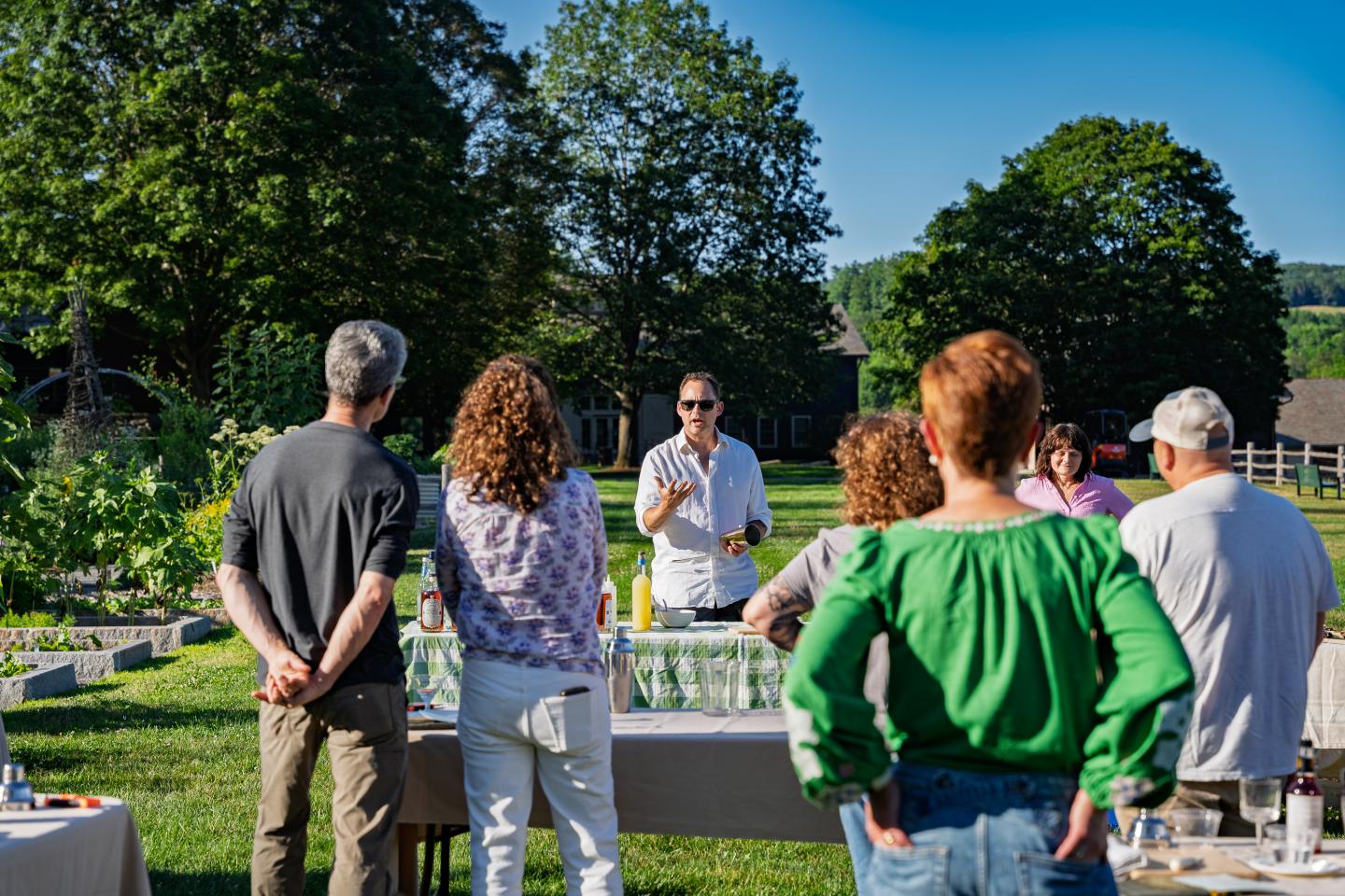 Group gathered outdoors, listening to a man speak under blue sky, surrounded by greenery.