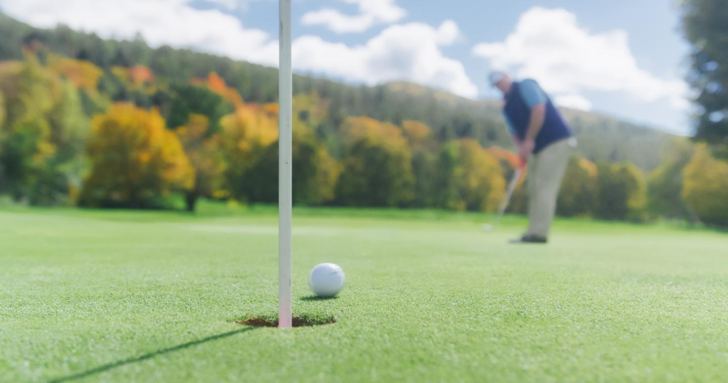 Golf ball near hole with player in background on a sunny day.