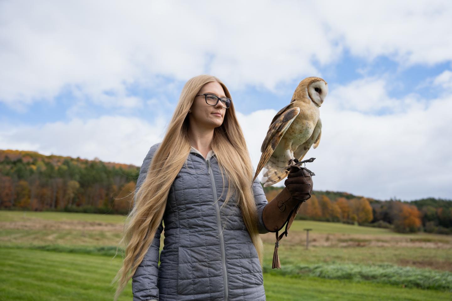 Woman with long hair, wearing glasses, holding an owl outdoors.