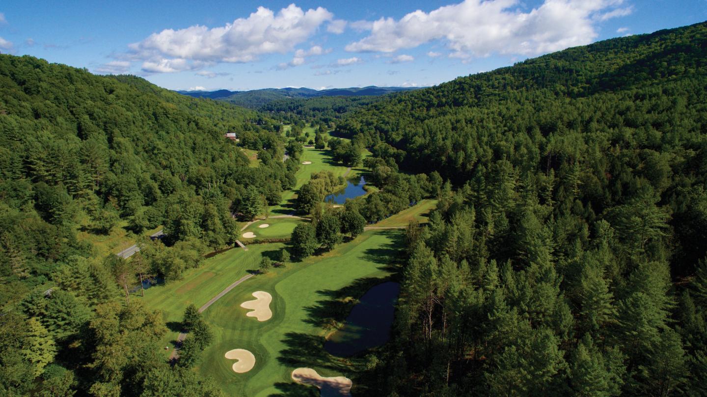 Golf course surrounded by forested hills under a blue sky with clouds.