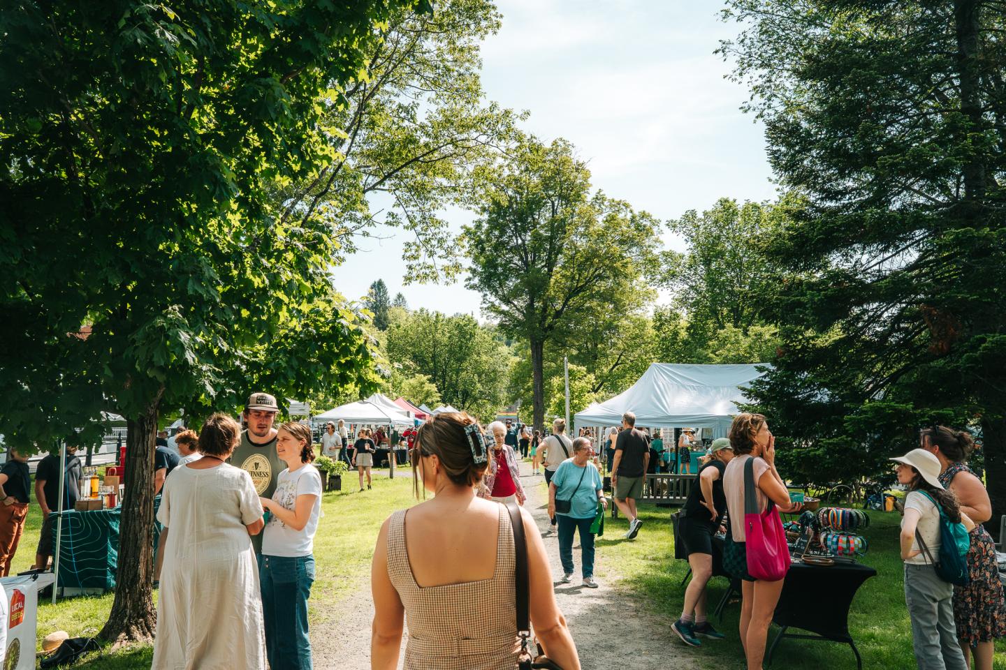 Outdoor market with people walking, surrounded by trees and tents.