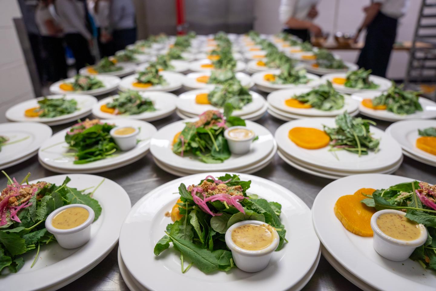 Plates of salad with dressing arranged on a long table in a kitchen setting.