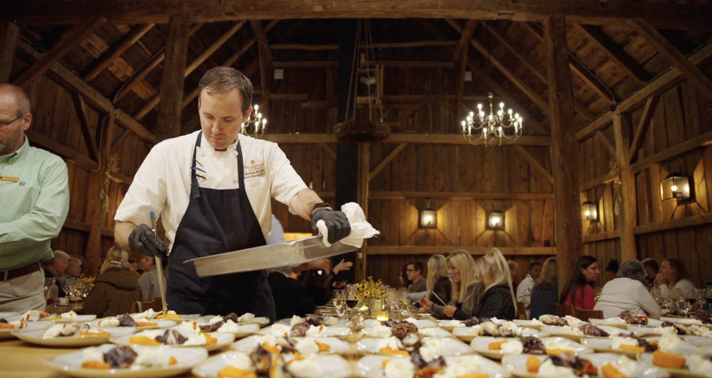 Chef plating dishes in a rustic wooden hall, with guests dining in the background.