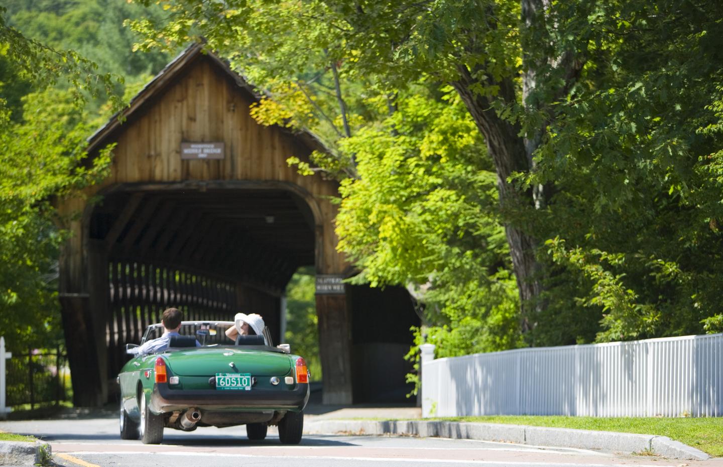 Green convertible driving through a wooden covered bridge surrounded by trees.